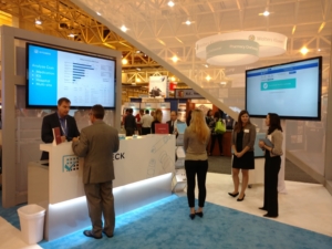 Trade show booth featuring an 80-inch monitor displaying data analytics, with attendees engaging in discussions and presentations in a professional setting.