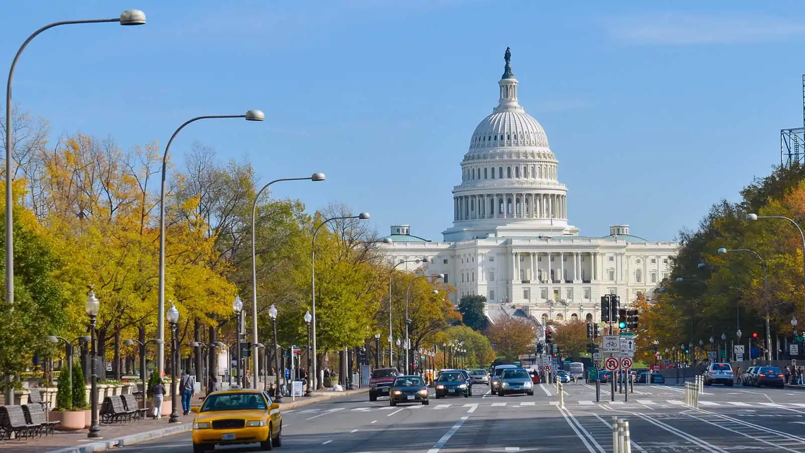 The photo captures a vibrant street scene in Washington, DC, with the iconic United States Capitol building prominently in the background. The foreground features a bustling avenue lined with trees, cars, and a yellow taxi, showcasing the dynamic and lively atmosphere of the city. The clear blue sky and autumn foliage add a picturesque quality to this quintessential view of the nation's capital.