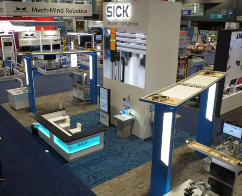 The photo shows a trade show booth for an exhibitor who are sensor intelligence experts. The booth has a big LED video wall with the company logo and tagline, “Sensor Intelligence” above a high def LED screen rental showing product images. The booth is modern with clean lines, bright lights and white and blue colours. At the front is a reception area with a desk branded with the company logo where visitors can be greeted and informed. To the right is a smaller workstation with more equipment or information. There are various high tech displays and interactive elements to show off the company’s technology. The other booths around the hall are robotics and automation focused with Mech-Mind Robotics next door. The whole atmosphere is high tech and professional for industry people looking for sensor and automation solutions.