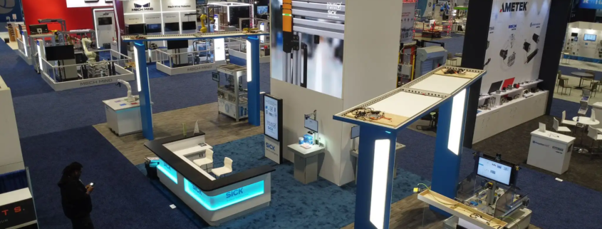 The photo shows a trade show booth for an exhibitor who are sensor intelligence experts. The booth has a big LED video wall with the company logo and tagline, “Sensor Intelligence” above a high def LED screen rental showing product images. The booth is modern with clean lines, bright lights and white and blue colours. At the front is a reception area with a desk branded with the company logo where visitors can be greeted and informed. To the right is a smaller workstation with more equipment or information. There are various high tech displays and interactive elements to show off the company’s technology. The other booths around the hall are robotics and automation focused with Mech-Mind Robotics next door. The whole atmosphere is high tech and professional for industry people looking for sensor and automation solutions.