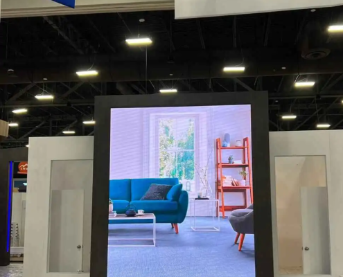 The video wall rental is prominent and high res in an exhibition or trade show environment. The LED screen rental shows a modern living room with a teal sofa, coffee table and window with natural light, bookshelf in the background. The display is framed in black to make the image pop.