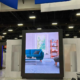 The video wall rental is prominent and high res in an exhibition or trade show environment. The LED screen rental shows a modern living room with a teal sofa, coffee table and window with natural light, bookshelf in the background. The display is framed in black to make the image pop.