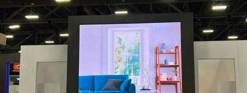 The video wall rental is prominent and high res in an exhibition or trade show environment. The LED screen rental shows a modern living room with a teal sofa, coffee table and window with natural light, bookshelf in the background. The display is framed in black to make the image pop.