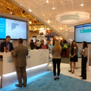 Trade show booth featuring a 70-inch monitor displaying data analytics, with attendees engaging in discussions and presentations in a professional setting.