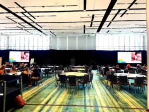 This is a conference room set up for an event. Round tables and chairs are arranged in a cozy layout. Big windows let in lots of natural light. Two big screens at the front are showing images and information. People are seated and chatting. A professional and collaborative space for meetings, conferences or seminars.