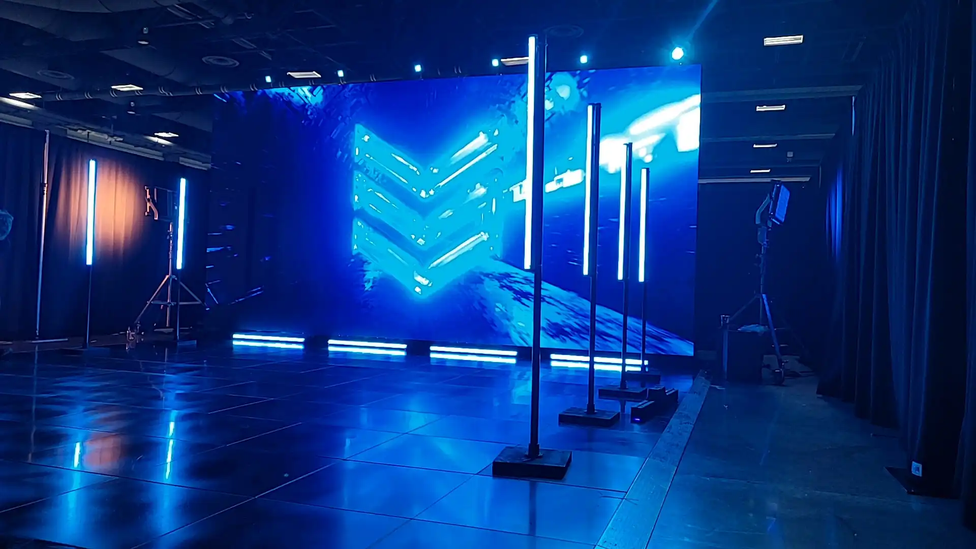 This photo depicts a modern audiovisual setup featuring a large digital screen with a vibrant blue abstract design and chevron patterns. The screen is surrounded by vertical LED light bars, casting a blue glow over the polished floor. The setting appears to be an event or conference space with black drapes, stage lighting equipment, and visible ceiling fixtures, creating a high-tech, immersive atmosphere.